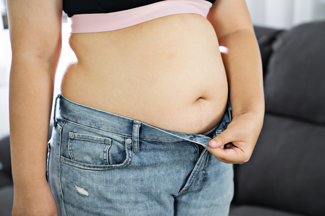 Close-up of a woman's midsection struggling to button tight jeans due to belly fat, linking to weight gain from alcohol.