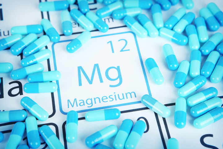 Blue capsules or supplements scattered around the chemical symbol for Magnesium (Mg) on a periodic table background.