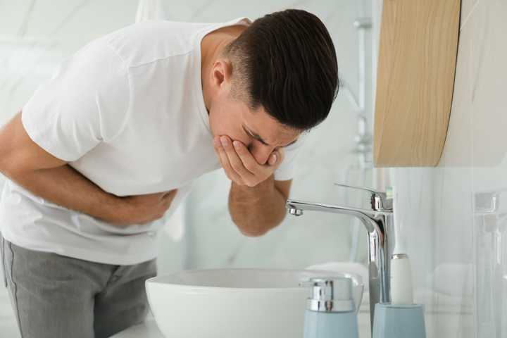A man bending over a bathroom sink, holding his hand to his mouth and stomach, showing the severe physical symptoms of hangover nausea.