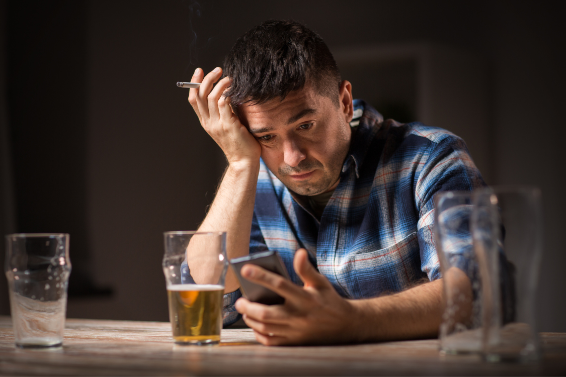 A man with a cigarette looking stressed while holding a phone next to empty and half-full glasses of beer, representing the "stress rebound" or 'hangxiety' effect after drinking alcohol.